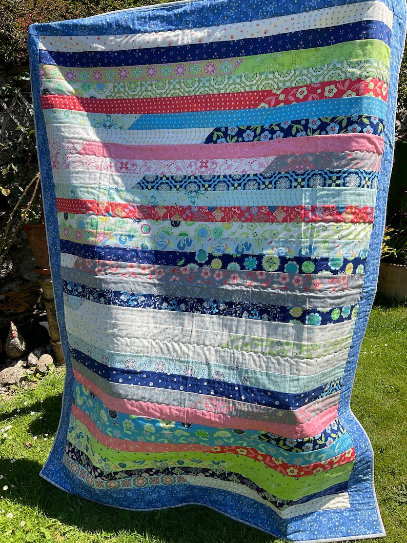 Op de afbeelding: Een blauw-wit gestreept quilt met een verscheidenheid aan kleurrijke stofstroken die in een horizontaal patroon aan elkaar zijn genaaid. De quilt is gemaakt met een verscheidenheid aan bloemen- en geometrische prints.