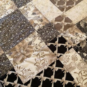 K&ouml;nnte beinhalten: Nahaufnahme eines Patchwork-Quilts mit einer Vielzahl von Mustern und Farben. Der Quilt zeigt Quadrate aus schwarzen, cremefarbenen und grauen Stoffen mit floralen, Schleifen- und geometrischen Designs. Die N&auml;hte sind sichtbar und verleihen Textur.