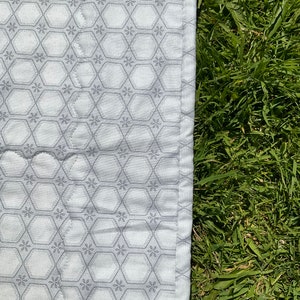 Op de afbeelding: Een witte stof met een grijs zeshoekig patroon en een witte rand. De stof ligt op een bed van groen gras.