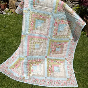 Puede incluir: Una colcha de patchwork con un patrón de bloques de cabaña de troncos en colores pastel suaves que incluyen azul claro, rosa pálido, verde salvia y crema. La tela muestra varios estampados florales, de cuadros y de lunares en una cuadrícula.