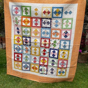 Puede incluir: Un edredón de patchwork hecho a mano con un borde blanco y un borde de cuadros naranja. El edredón presenta una cuadrícula de cuadrados coloridos con varios patrones y colores, incluyendo rojo, azul, amarillo y verde. El edredón se muestra al aire libre.