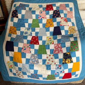 May include: A patchwork quilt with a blue and white chequered design. The quilt incorporates a variety of colourful fabric squares, including red, yellow, green, and floral patterns. It is edged with a solid blue fabric.