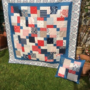 Op de afbeelding: Een patchworkdeken in een blauw, rood en wit kleurenschema. De deken is gemaakt van vierkanten van verschillende stoffen en is afgewerkt met een blauwe en witte geprinte stof. Een bijpassend kussen is ook te zien.