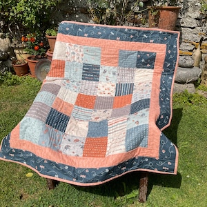 Könnte beinhalten: Ein Patchwork-Quilt mit quadratischen Mustern in Blau-, Pfirsich- und Grautönen. Der Quilt hat einen dunkelblauen Rand mit Libellenmuster und einen pfirsichfarbenen Innenrand. Der Quilt liegt auf einer Holzbank. Maße nicht sichtbar.