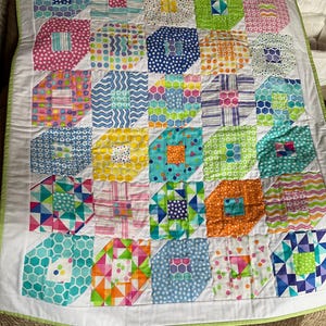 May include: A colorful patchwork quilt with a white border and green trim. The quilt features a repeating pattern of geometric shapes in various colors and patterns, including polka dots, stripes, and hexagons. The quilt is likely handmade.