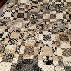 K&ouml;nnte beinhalten: Eine Patchwork-Decke in Schwarz-, Wei&szlig;- und Beiget&ouml;nen. Die Decke besteht aus zahlreichen Quadraten mit verschiedenen Mustern, darunter florale Designs, geometrische Formen und Schleifenmotive. Die Decke liegt auf einem Bett.