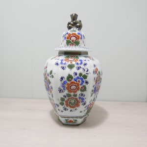 May include: A white ceramic jar with a lid featuring a floral design in shades of blue, orange, and green. The lid has a small, brown, animal-shaped finial.