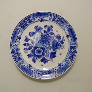 Könnte beinhalten: Ein runder, weißer Keramikteller mit blauem Blumendesign. Der Teller zeigt einen zentralen Blumenstrauß aus blauen Blumen und einen detaillierten blauen Rand. Das Design erinnert an Delfter Ware.