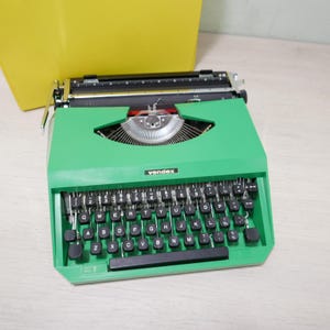 May include: A vintage green Vendex typewriter with black keys and a silver carriage. The keyboard features white letter and number keys. The word "vendex" is printed on the front of the typewriter. A yellow carrying case is visible in the background.