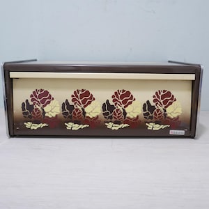 May include: A brown metal bread box with a floral design in shades of burgundy, cream, and yellow. The box has a hinged lid and chrome accents. The brand name "brabantia" is visible on the lower right corner.