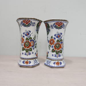 Könnte beinhalten: Zwei weiße Keramikvasen mit blauen, orangefarbenen und grünen Blumendesigns. Die Vasen haben einen goldenen Rand und eine sechseckige Form.