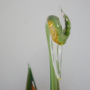May include: Close-up of a green glass swan sculpture with a clear glass beak. The swan's head is tilted to the side, and the beak is slightly open.