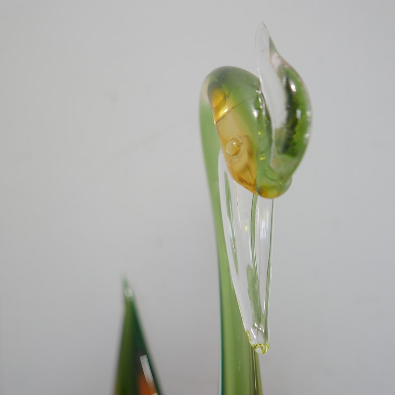 May include: Close-up of a green glass swan sculpture with a clear glass beak. The swan's head is tilted to the side, and the beak is slightly open.