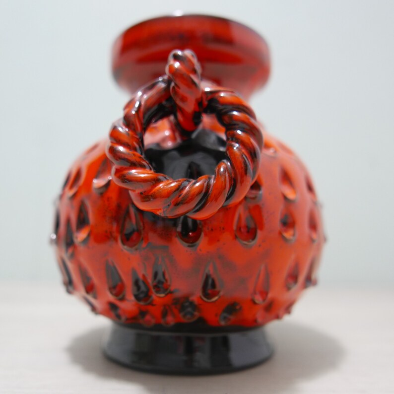 May include: A red and black ceramic vase with a rope-like handle. The vase has a textured surface with small, raised dots.