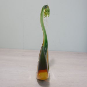 May include: A green and amber glass swan figurine with a long neck and a curved body. The swan is standing on a white surface.