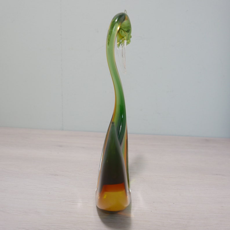 May include: A green and amber glass swan figurine with a long neck and a curved body. The swan is standing on a white surface.