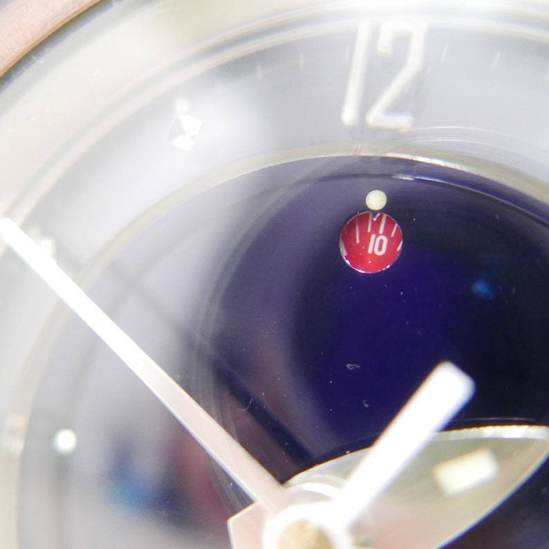 May include: Close-up of a clock face with a white background and silver hands. The clock face has a blue center with a red circle containing the number "10" in white.