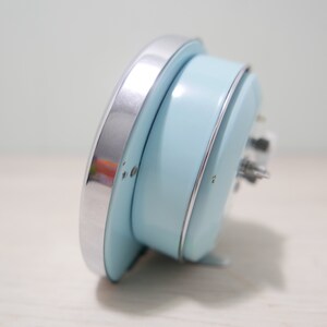 May include: A light blue alarm clock with a silver rim. The clock is lying on its side, showing the back of the clock.
