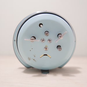 May include: A light blue alarm clock with silver accents and a silver knob on the side. The clock has a white face with black numbers and hands.