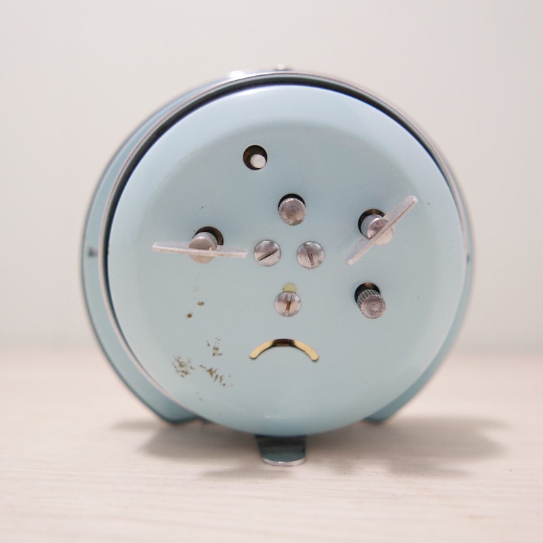May include: A light blue alarm clock with silver accents and a silver knob on the side. The clock has a white face with black numbers and hands.