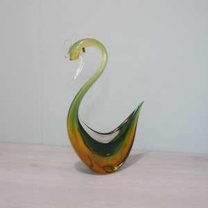 May include: A green and amber glass sculpture of a stylized bird. The bird has a long neck and a curved body.