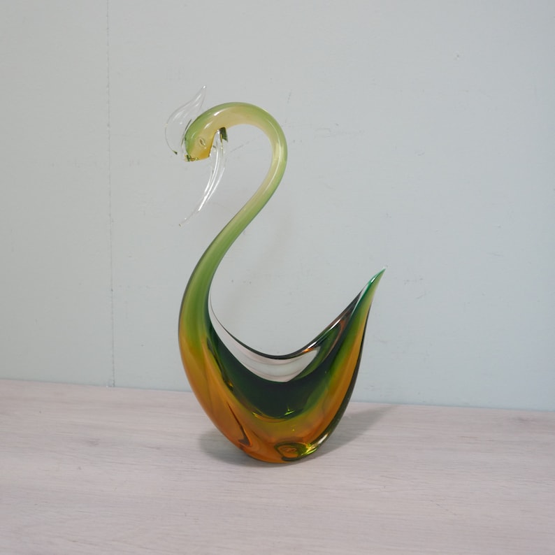May include: A green and amber glass sculpture of a stylized bird. The bird has a long neck and a curved body.