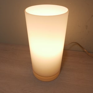 K&ouml;nnte beinhalten: Eine wei&szlig;e, mattierte Tischlampe aus Glas mit runder Basis. Die Lampe ist eingeschaltet und strahlt ein warmes, wei&szlig;es Licht ab.