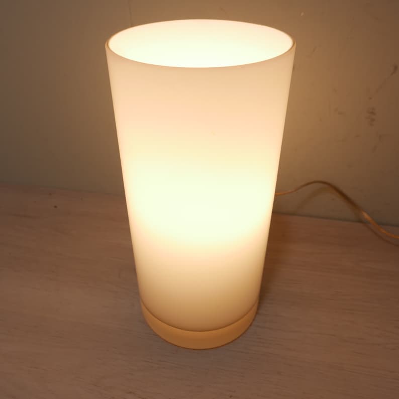 K&ouml;nnte beinhalten: Eine wei&szlig;e, mattierte Tischlampe aus Glas mit runder Basis. Die Lampe ist eingeschaltet und strahlt ein warmes, wei&szlig;es Licht ab.