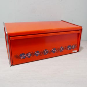 May include: A vintage orange bread box with a floral design. The rectangular box has a hinged lid and a silver trim. The bottom of the box features a row of blue and purple flowers. A retro kitchen storage item.