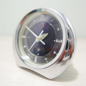 May include: A silver alarm clock with a black face and white numbers. The clock has a dark blue dial with a silver hand and a red dot at the 12 o'clock position.