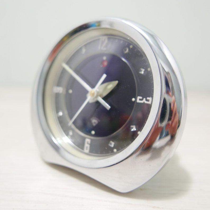 May include: A silver alarm clock with a black face and white numbers. The clock has a dark blue dial with a silver hand and a red dot at the 12 o'clock position.