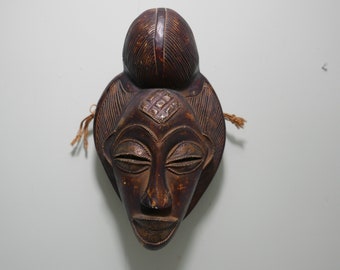 Pode incluir: Uma máscara de madeira castanho-escura com um rosto esculpido e um coque no topo da cabeça. A máscara tem um padrão geométrico na testa e os olhos fechados.