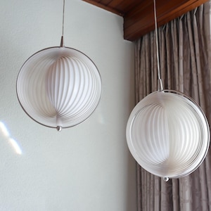 Könnte beinhalten: Zwei kugelförmige Pendelleuchten mit weißem, geripptem Innenraum und einem silberfarbenen Metallrahmen. Die Leuchten hängen an dünnen Drähten und schaffen eine moderne und minimalistische Ästhetik. Der Hintergrund umfasst eine weiße Wand und einen Vorhang.