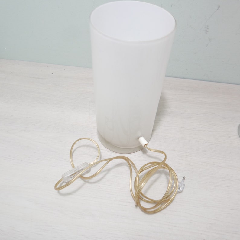 K&ouml;nnte beinhalten: Eine wei&szlig;e, mattierte Tischlampe aus Glas mit einem goldenen Kabel. Die Lampe hat ein schlichtes, zylindrisches Design.