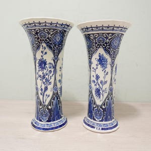 Może przedstawiać: Dwie pasujące do siebie ceramiczne wazony z rozszerzanym brzegiem i podstawą, posiadające białe tło ozdobione misternymi niebieskimi wzorami kwiatowymi oraz dekoracyjnymi obwódkami, które otaczają cylindryczny korpus każdego naczynia.
