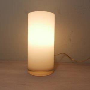 K&ouml;nnte beinhalten: Eine wei&szlig;e, mattierte Tischlampe aus Glas mit runder Basis und zylindrischem Schirm. Die Lampe ist eingeschaltet und strahlt ein warmes, gelbes Licht aus.