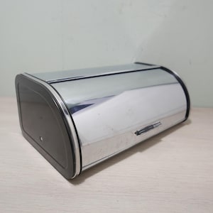 May include: A chrome bread box with a black end panel. The bread box has a curved top and a small handle. The bread box is sitting on a light-colored wooden surface.