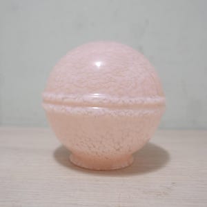 Op de afbeelding: Een lichtroze glazen lampenkap met een gespikkeld patroon. De ronde kap heeft een duidelijke band in het midden en een kleine voet. Het glas heeft een glanzende afwerking. De lampenkap is waarschijnlijk vintage of antiek.