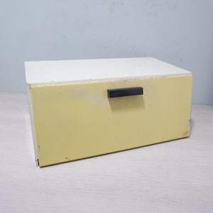 May include: A rectangular storage box with a white top and a pale yellow body. It has a black handle and appears to be made of metal. The box is sitting on a light-colored surface.