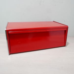 Puede incluir: Una panera roja brillante con forma rectangular y puerta frontal abatible. La panera tiene una superficie lisa y brillante y un ribete plateado en los bordes. La panera está diseñada para guardar pan y productos de panadería.