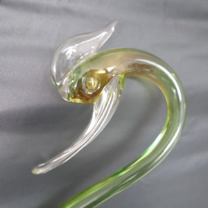 May include: A green glass sculpture of a bird with a clear glass head and wings. The bird's head is tilted to the side and its beak is open.