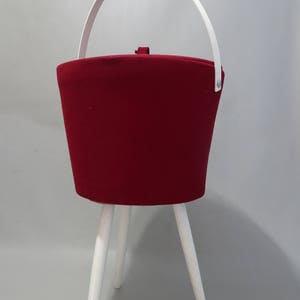 May include: A red fabric storage ottoman with a white handle and white wooden legs.