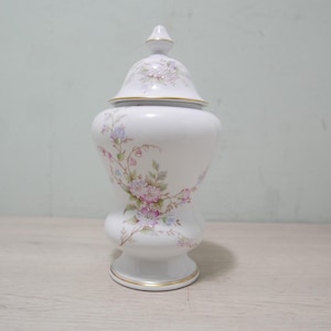 May include: A white porcelain jar with a lid, decorated with floral patterns in pink, purple, and green. The jar has a bulbous body, a pedestal base, and gold trim. The lid is topped with a small finial.