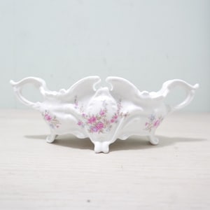 Puede incluir: Un centro de mesa de porcelana blanca con bordes festoneados ornamentados y dos asas laterales. El exterior está decorado con delicados motivos florales en tonos rosa y azul, y el recipiente se apoya sobre cuatro pequeños pies curvos.