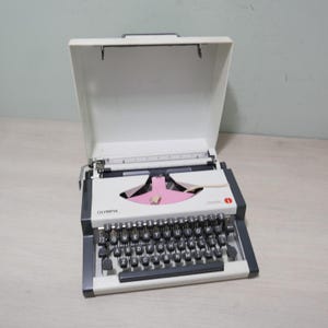 May include: Olympia Traveler typewriter with a white body, black keys, and a pink ribbon. The typewriter is in its open, white carrying case. The Olympia logo is visible on the front of the typewriter.