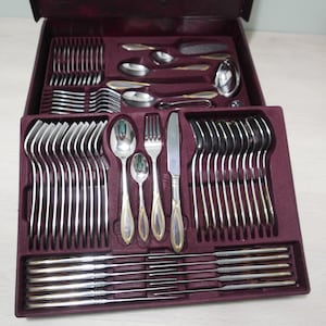 Könnte beinhalten: Ein Set silbernes Besteck in einem burgunderfarbenen, samtenen Etui. Das Besteck umfasst Gabeln, Messer, Löffel und Teelöffel. Das Set ist in Reihen angeordnet und bereit für ein formelles Abendessen.