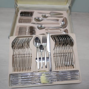Könnte beinhalten: Ein Set aus Edelstahlbesteck in einem weißen Etui. Das Besteck ist in Reihen angeordnet und umfasst Gabeln, Messer, Löffel und Servierbesteck. Das Etui trägt ein Etikett mit der Aufschrift "Classic International Stainless Steel"