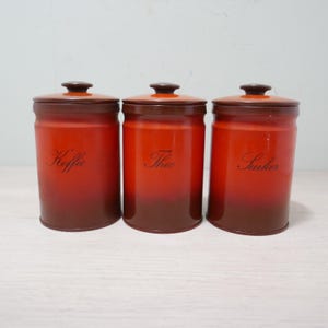 May include: Three vintage red and brown canisters with lids. The canisters are labeled "Koffie", "Thee", and "Suiker" in elegant script. The color gradient transitions from a deep brown at the base to a vibrant red at the top.