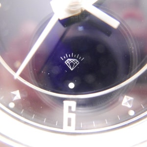 May include: A black and white clock face with a diamond symbol in the center. The clock face is surrounded by a silver ring with black markings. The clock face is set against a white background.