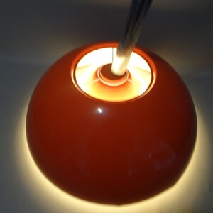 May include: A bright orange lampshade with a white interior and a silver metal stem. The lampshade is lit and casting a warm glow.
